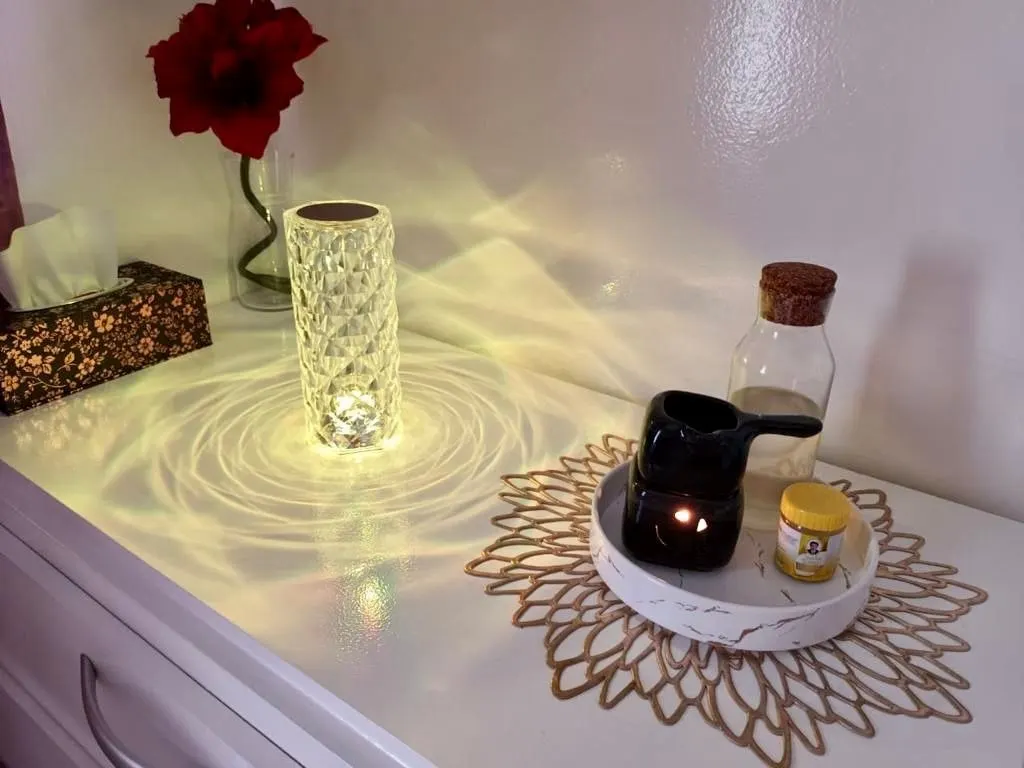 Sur une commode blanche : une lampe en cristal allumée, un brûle-parfum noir et une rose rouge sur un napperon doré