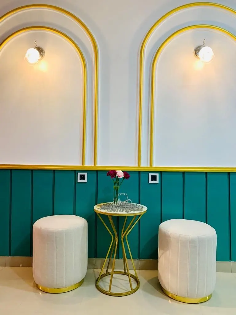 Le salon : lambris bleu canard, deux arches cernées d’or avec appliques, deux poufs blancs et une table dorée