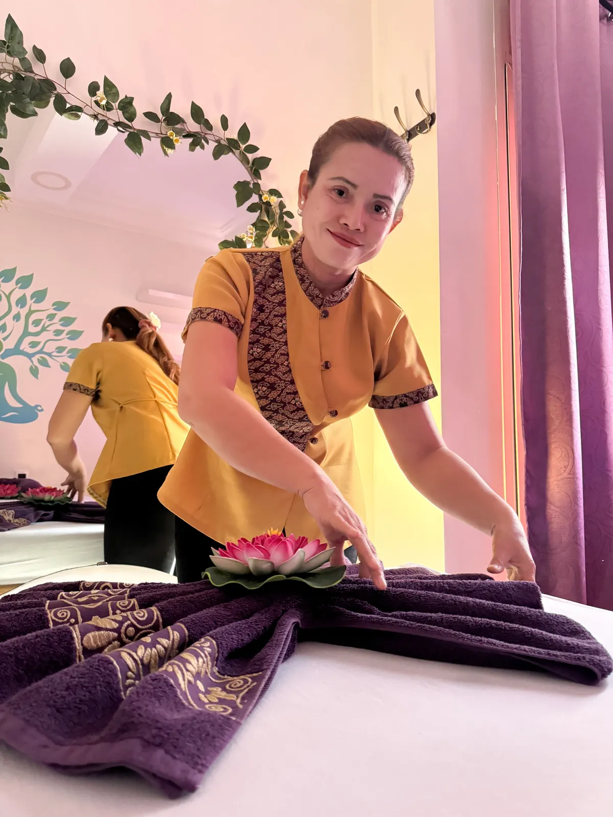 Une masseuse en tunique safran pose un lotus rose sur une serviette violette